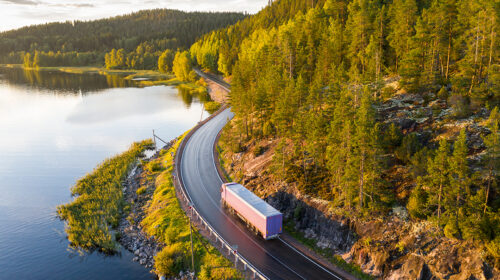 Aerial view of semi truck with cargo trailer on road curve at lake shore with green pine forest. Transportation background. Beautiful nature landscape at sunset in Republic of Karelia, Russia