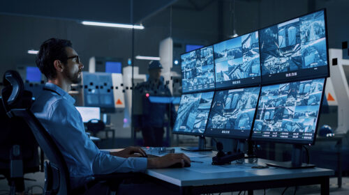 Industry 4.0 Modern Factory: Security Operator Controls Proper Functioning of Workshop Production Line, Uses Computer with Screens Showing Surveillance Camera Feed. High-Tech Security